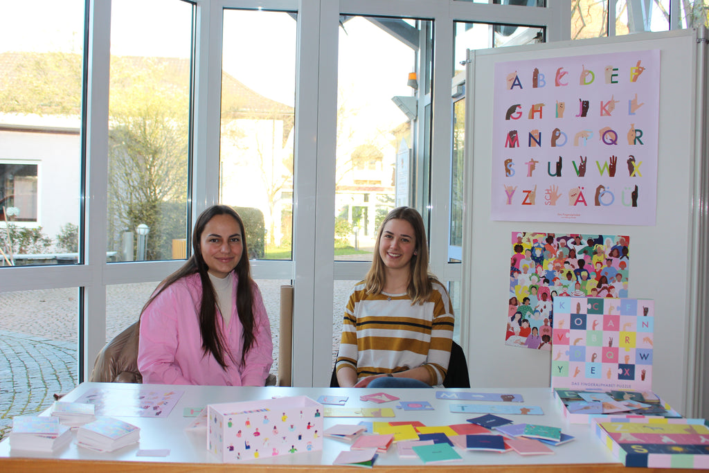 Katja und Emilia sind zu sehen, wie sie einen Workshop für Gebärden mit den Produkten von talking hands geben. Im Hintergrund sind die Gebärden-Daumenkinos, das Fingeralphabet Puzzle und die Poster zu sehen.